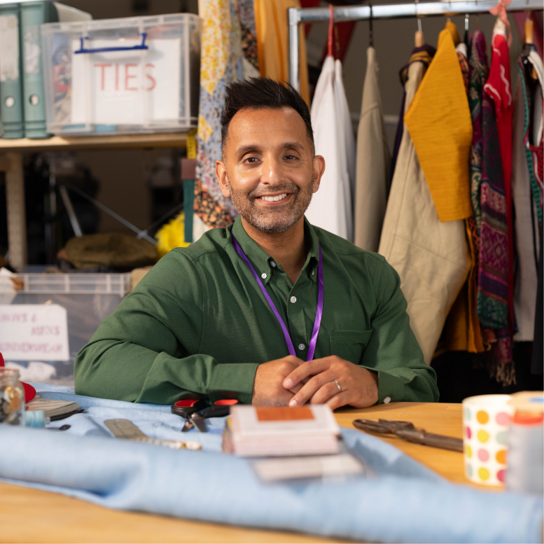 ITV Celebrity Doctor, Dr Amir Khan, smiles at the camera. He is sitting inside at a table wearing a smart shirt and a lanyard. He is surrounded by textiles as if he is about to upcycle some clothes. On the table in front of him is some material and scissors. Behind him there are labelled boxes with more textiles and a hanger filled with clothes. 